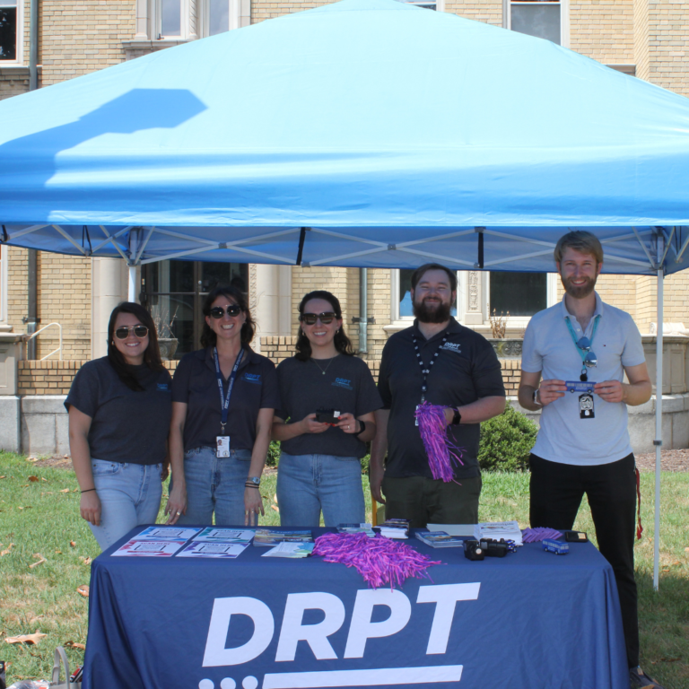 DRPT کا عملہ نیلے خیمے کے نیچے DRPT برانڈڈ ٹیبل پر پروموشنل آئٹمز رکھے ہوئے ہے۔